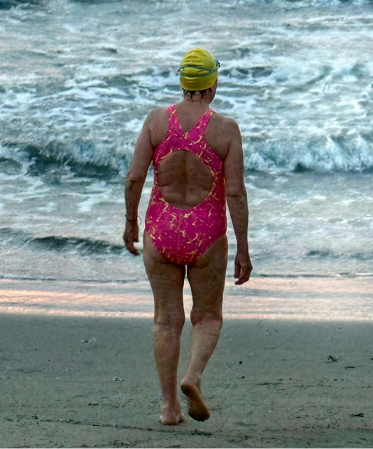 Person in a pink swimsuit and yellow swim cap walking on a beach with waves in the background