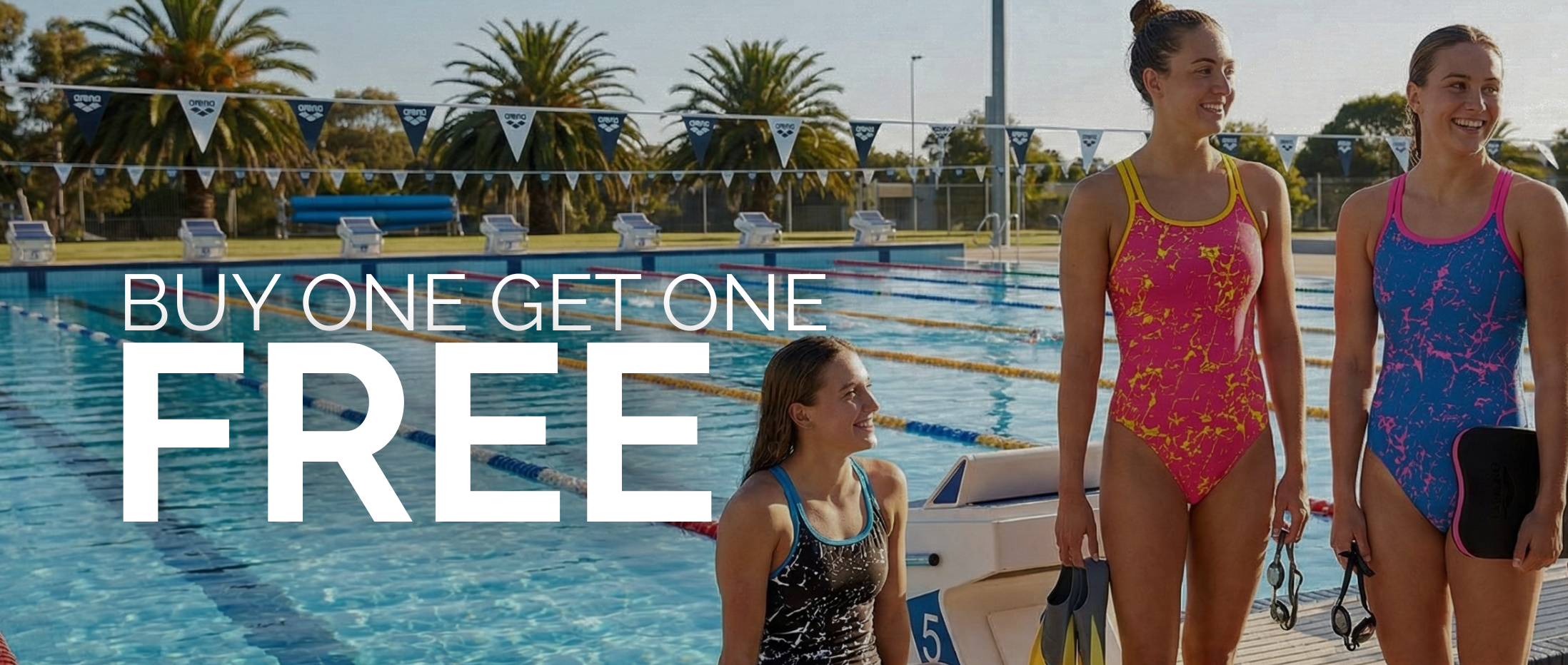 Four women in swimsuits standing and sitting by an outdoor swimming pool, with large overlaid text reading 'BUY ONE GET ONE FREE'. The scene includes pool lanes, palm trees, and a bright sky, so this is Lifestyle rather than Product/Packaging.